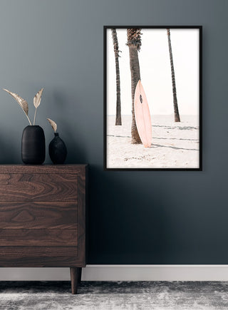 Framed print of a pink surfboard leaning against a palm tree on a sandy beach, displayed above a dark dresser.