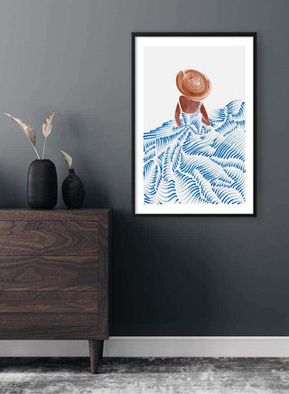 Art print of a woman in a summer dress and hat, standing among blue abstract waves, framed on a dark wall.