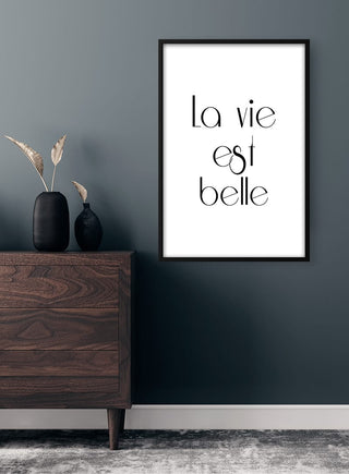 La Vie est Belle poster framed on a dark wall above a wooden dresser with decorative vases and dried plants.