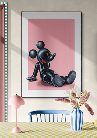 Framed print of a black cartoon figure lounging on pink, displayed above a table with flowers, for Kawsey Relax - Daniel Leo.