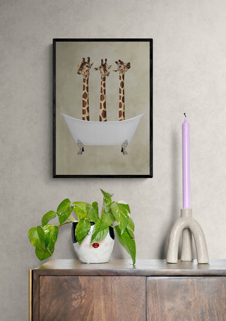 Framed print of three giraffes in a bathtub by Coco de Paris, displayed above a cabinet with decor.